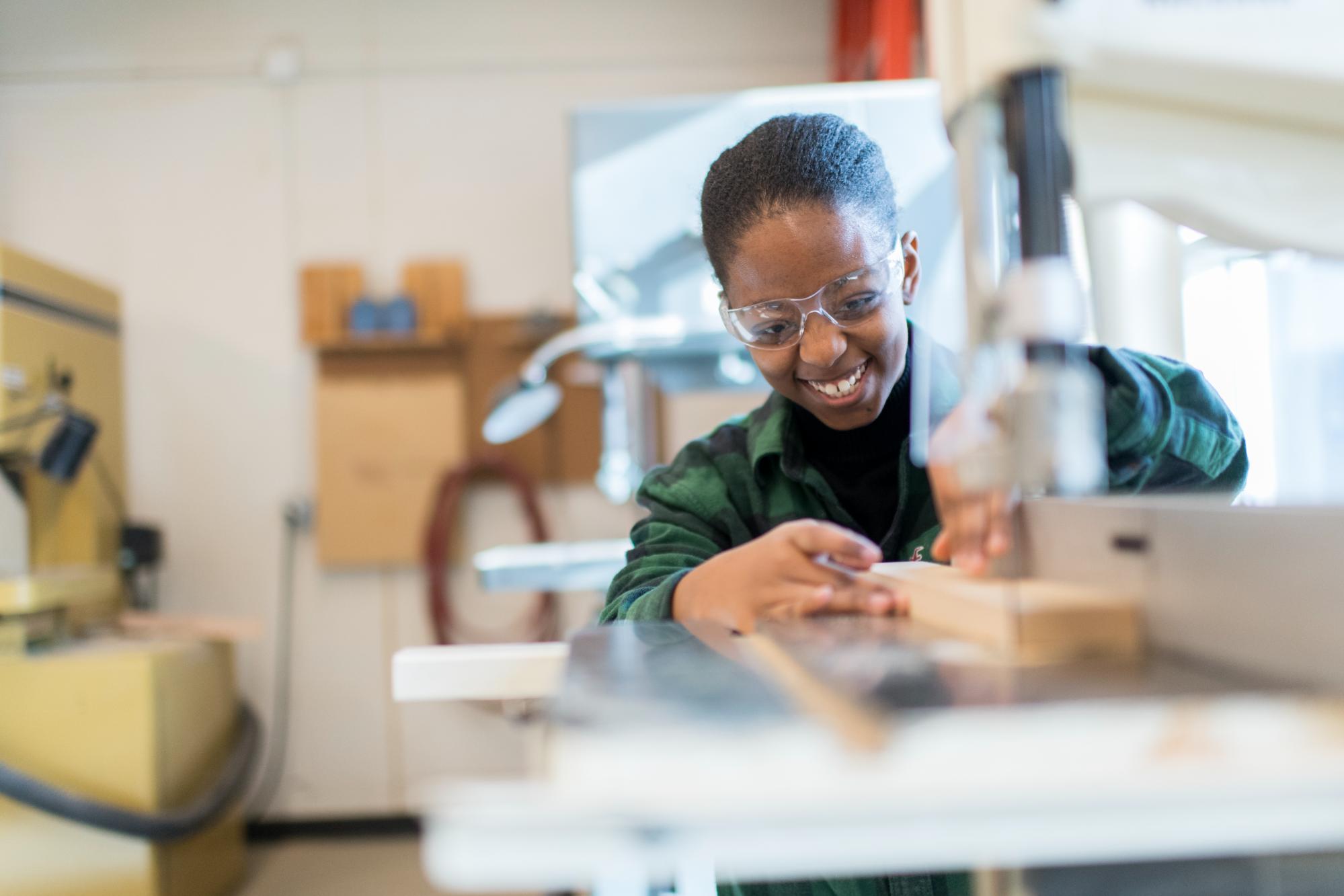 A young woman wearing safety goggles smiling while using a machine to cut wood